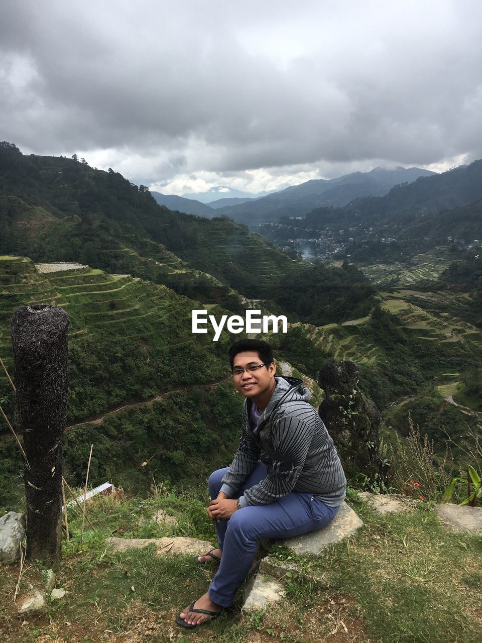 Portrait of young man sitting on mountain against cloudy sky