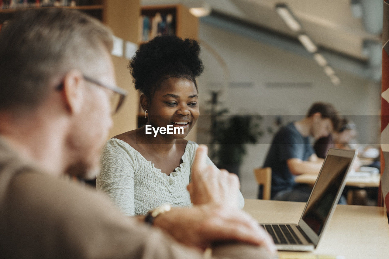 Smiling student discussing with professor over laptop in library at university