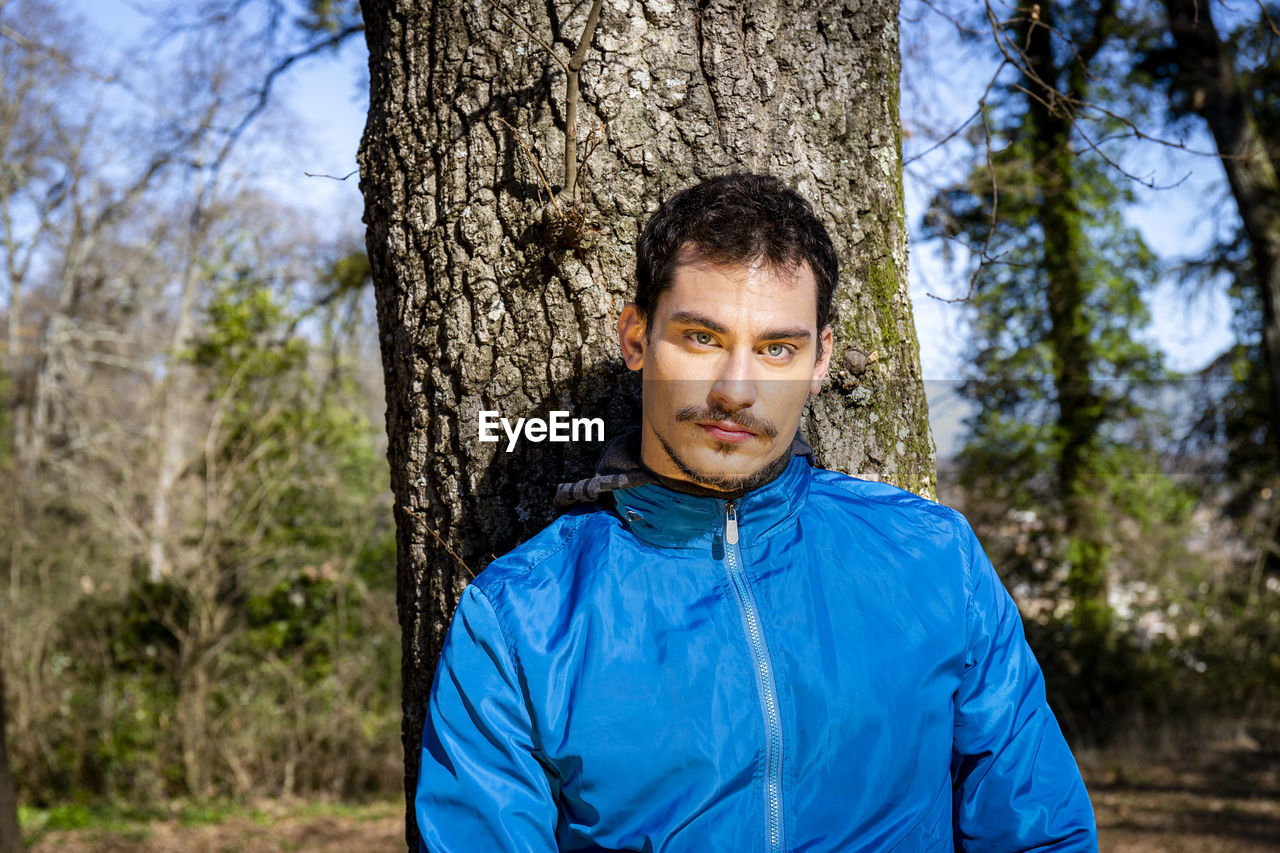 Beautiful man with blue eyes. the man is training in the woods and is leaning against an oak tree.