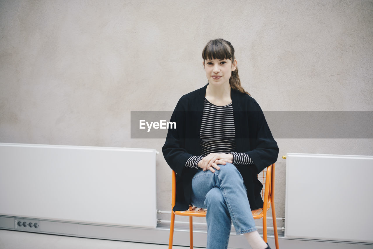 Portrait of confident female computer programmer sitting on chair against beige wall in office