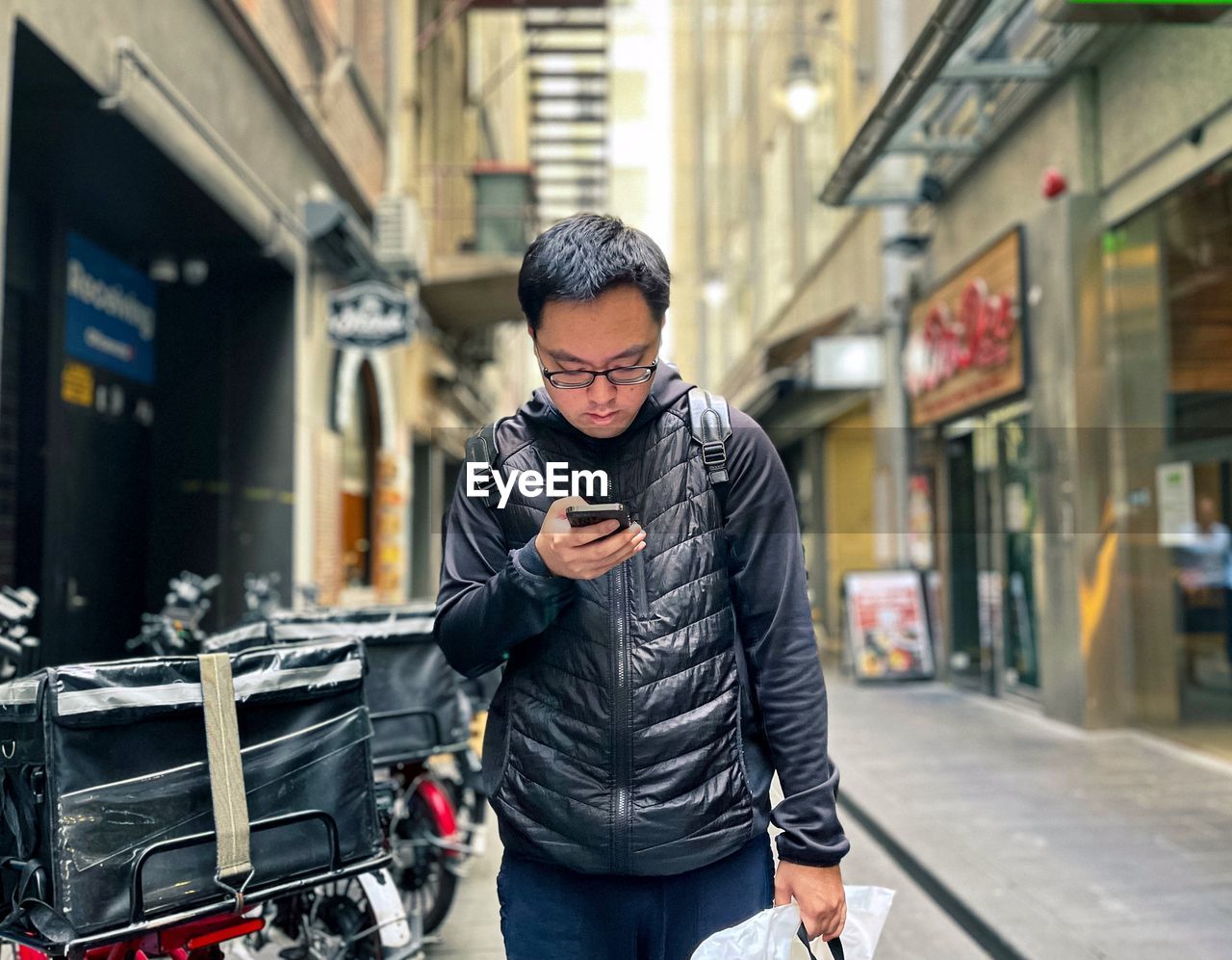 Portrait of young asian man using smart phone in laneway against buildings in the city.