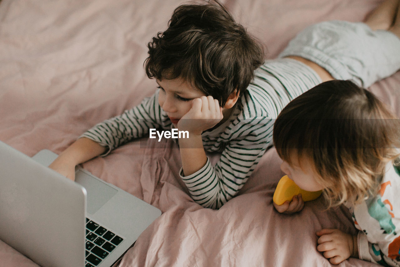 Little brother and sister lie in the bedroom on the bed and look at the laptop. high quality photo