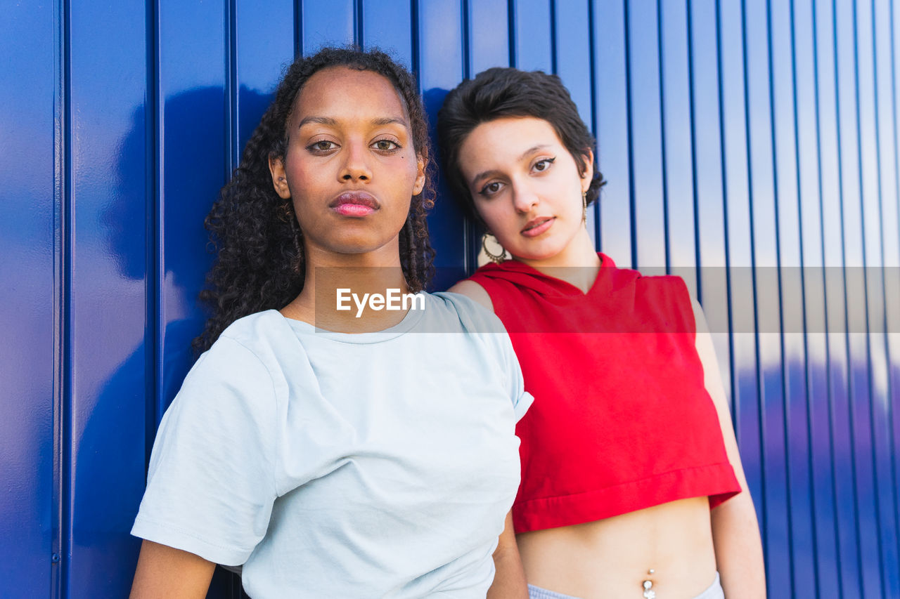 Serious multiracial young female friends in casual clothes looking at camera while standing together against blue background on street of city