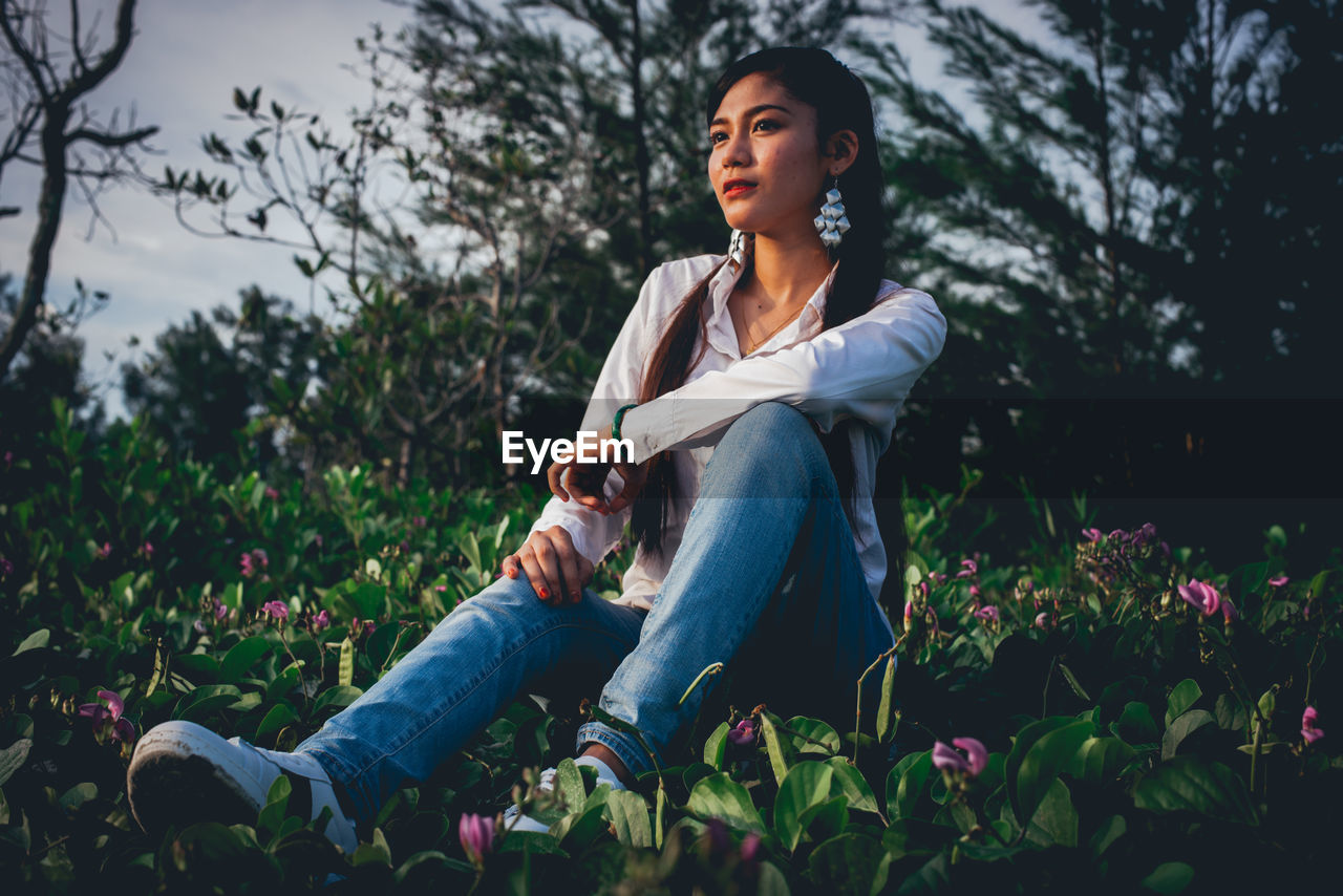 Thoughtful young woman looking away while sitting on field against trees