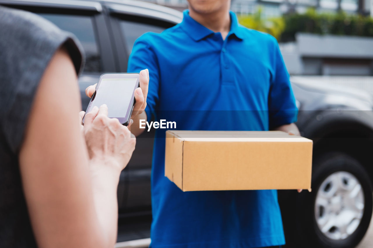 Midsection of woman holding mobile phone while delivery man holding box outdoors