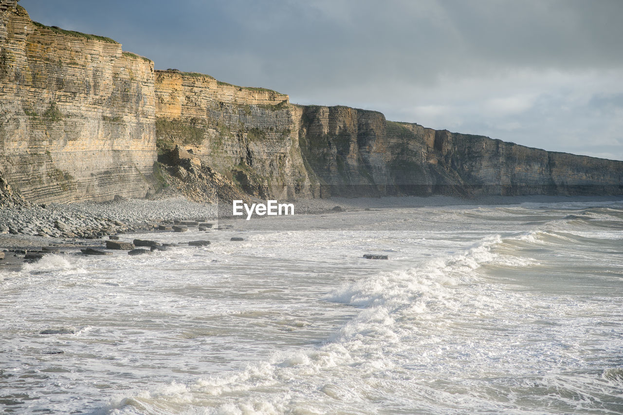 Scenic view of sea waves splashing on cliff against cloudy sky