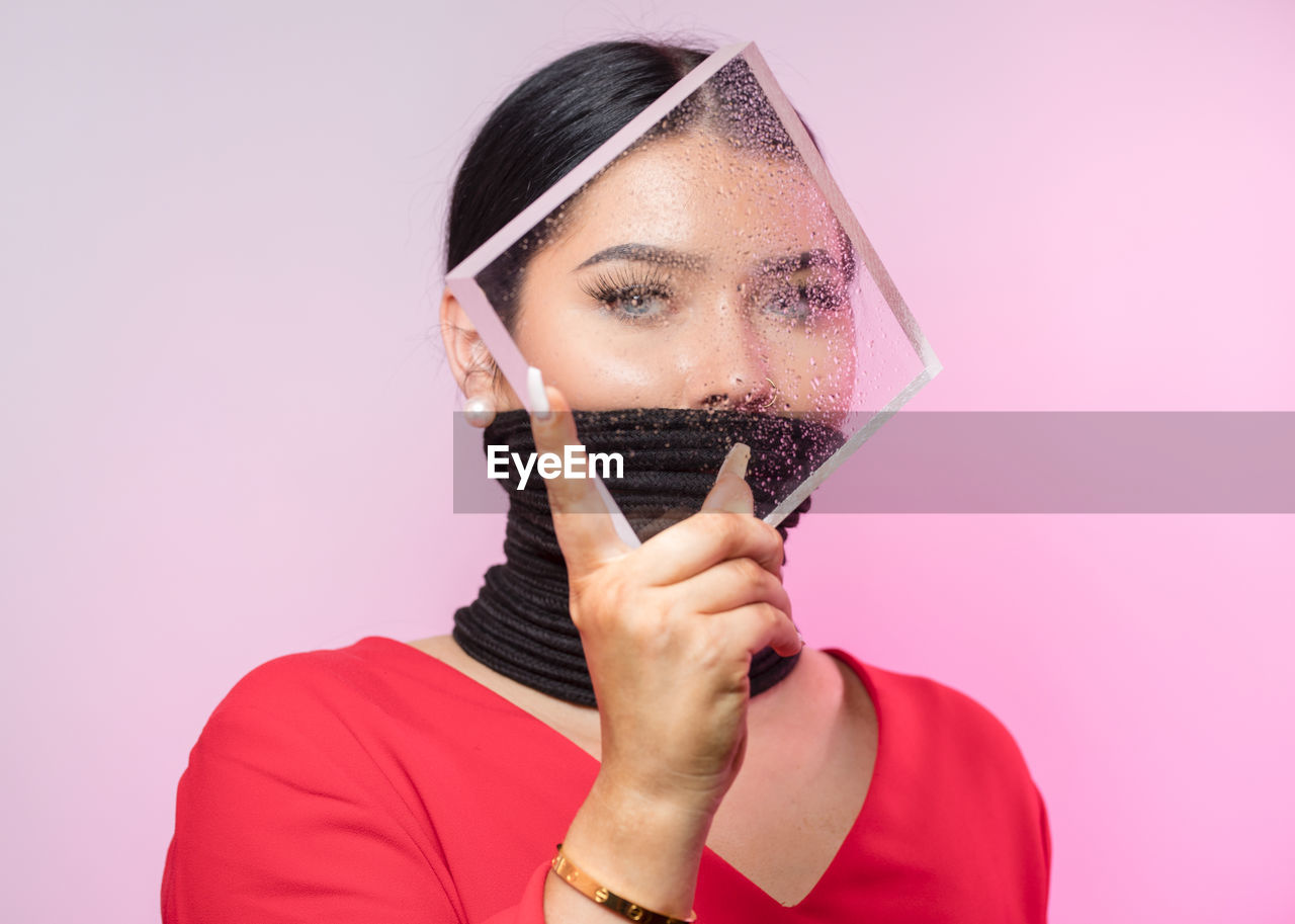 Portrait of young woman with tied mouth holding wet glass against pink background