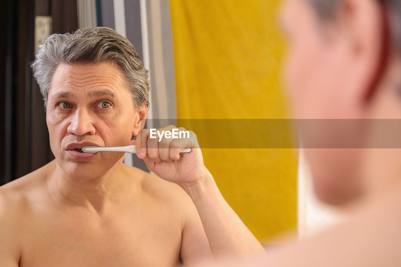 An adult gray-haired man brushes his teeth with a toothbrush and looks in the mirror