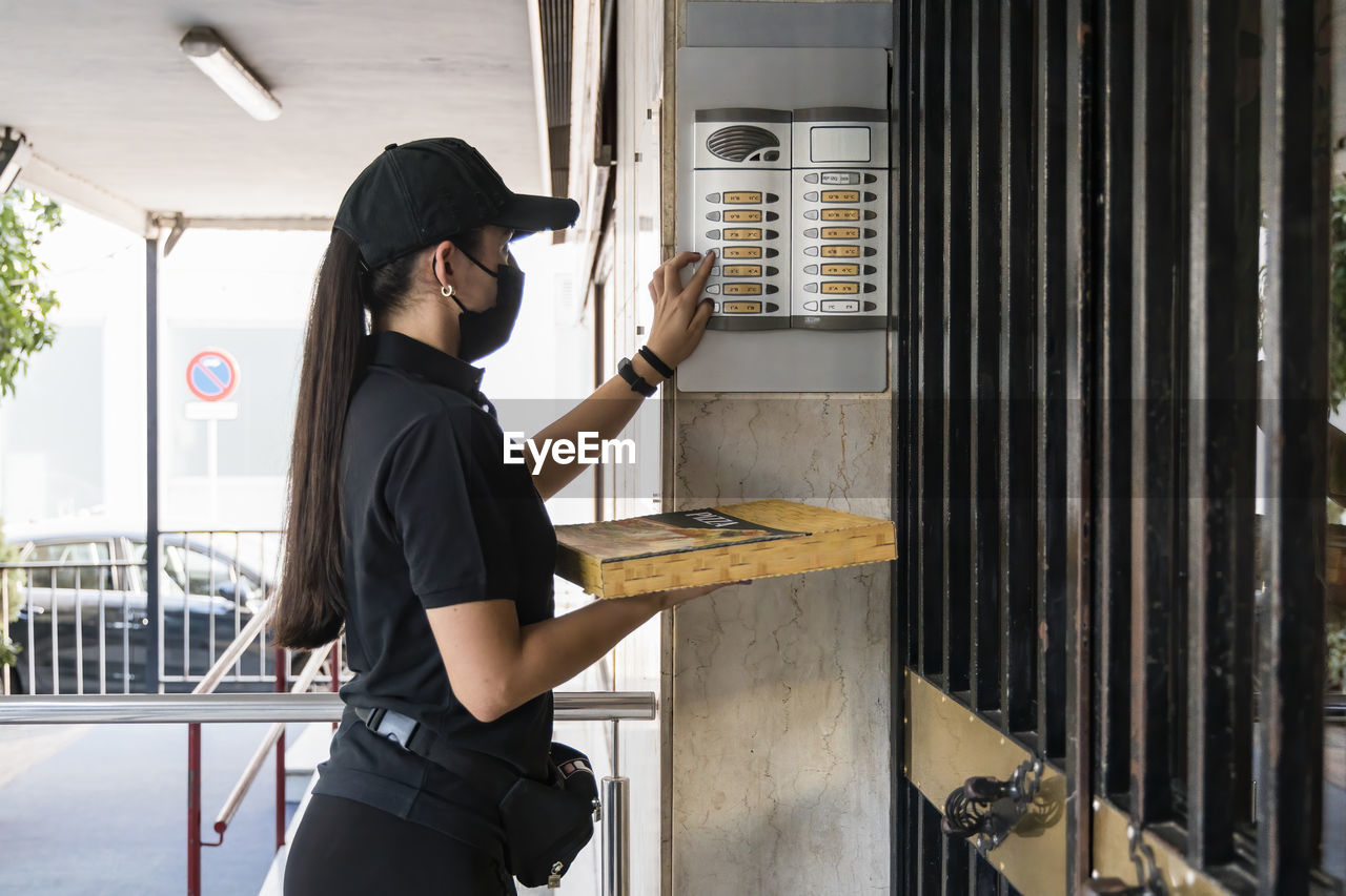 Young pizza delivery woman holding box while ringing doorbell during pandemic