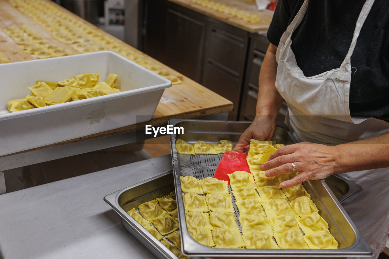food, food and drink, italian food, freshness, adult, occupation, pasta, indoors, business, one person, kitchen, fast food, meal, dish, business finance and industry, baked, cuisine, midsection, domestic room, hand, working, yellow, food and drink establishment, holding, preparing food, chef, healthy eating, small business, container, food and drink industry, lunch, standing, women, cooking