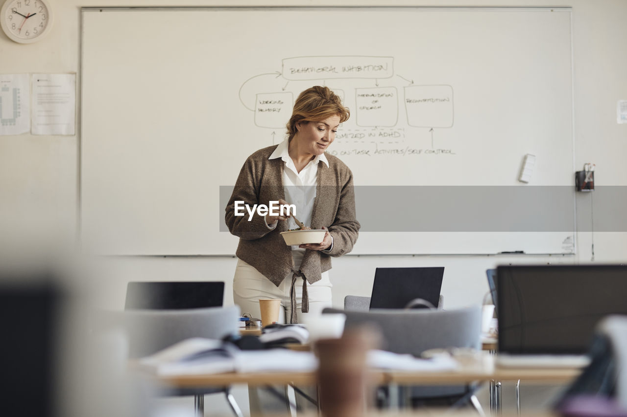 Female teacher holding lunch box while looking at laptop in classroom