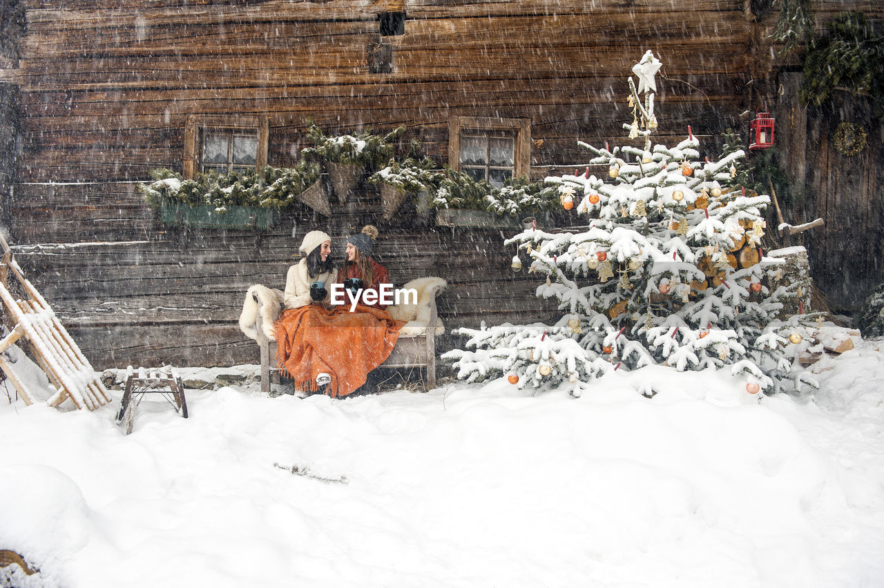 Friends sitting on bench by christmas tree in front of mountain hut