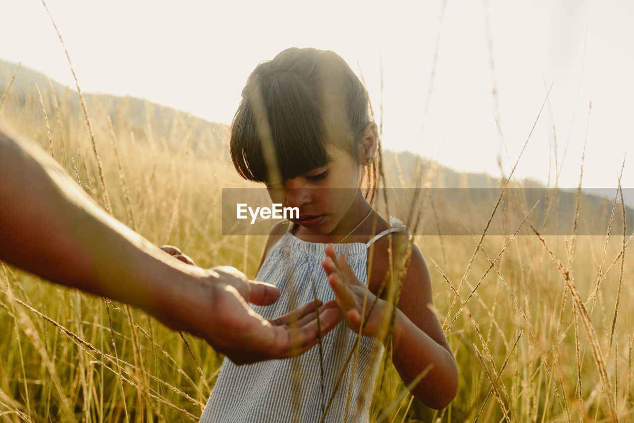 Unrecognizable crop adult parent extending hand to little girl against blurred of green meadow in daylight