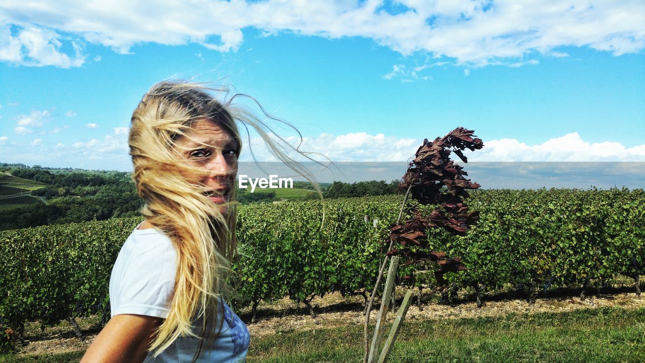 Portrait of woman with tousled hair standing at farm against sky