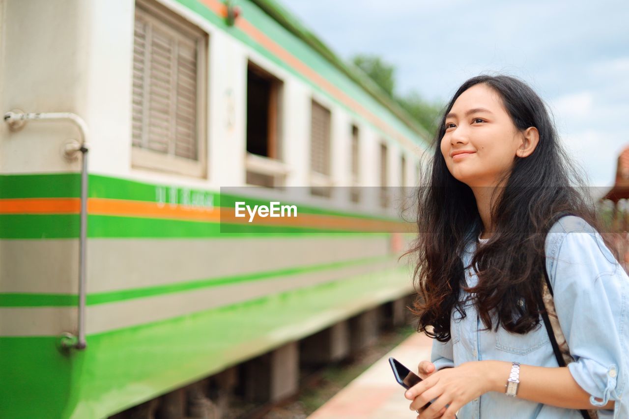 YOUNG WOMAN USING PHONE WHILE STANDING BY TRAIN