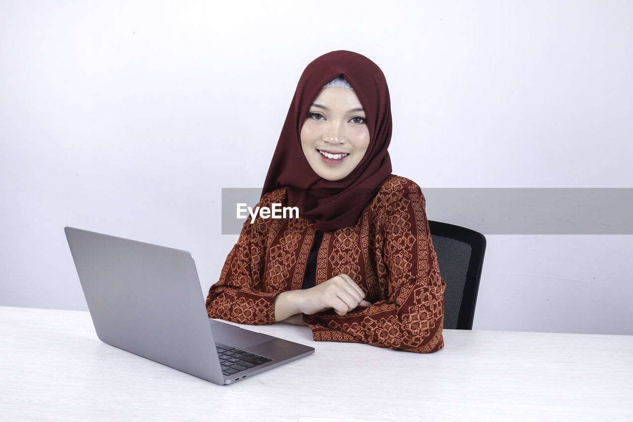 PORTRAIT OF SMILING YOUNG WOMAN USING SMART PHONE IN LAPTOP
