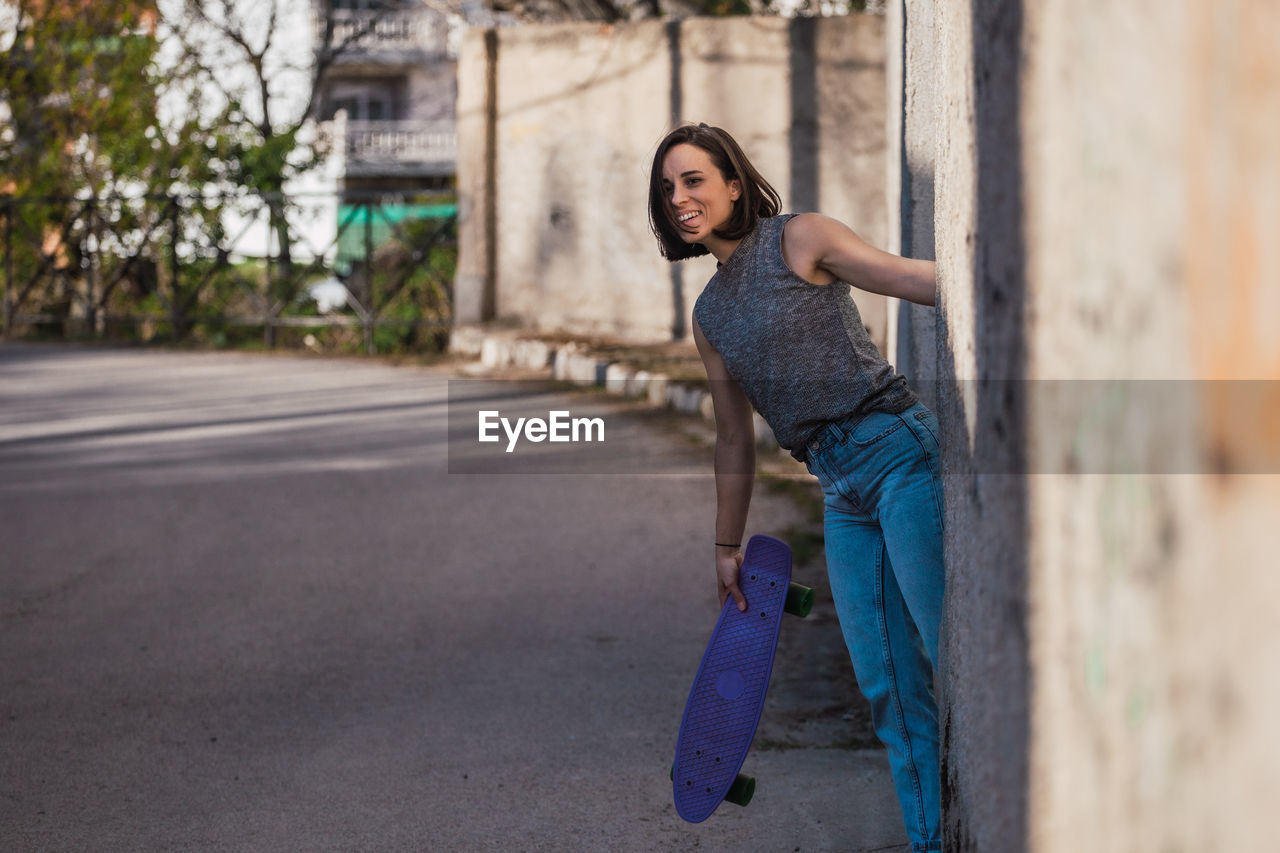 Portrait of smiling young woman holding skateboard by road