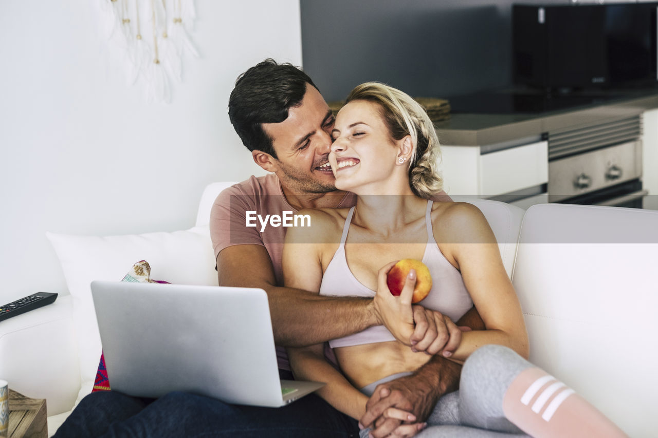 Smiling man and woman kissing while using laptop on sofa at home