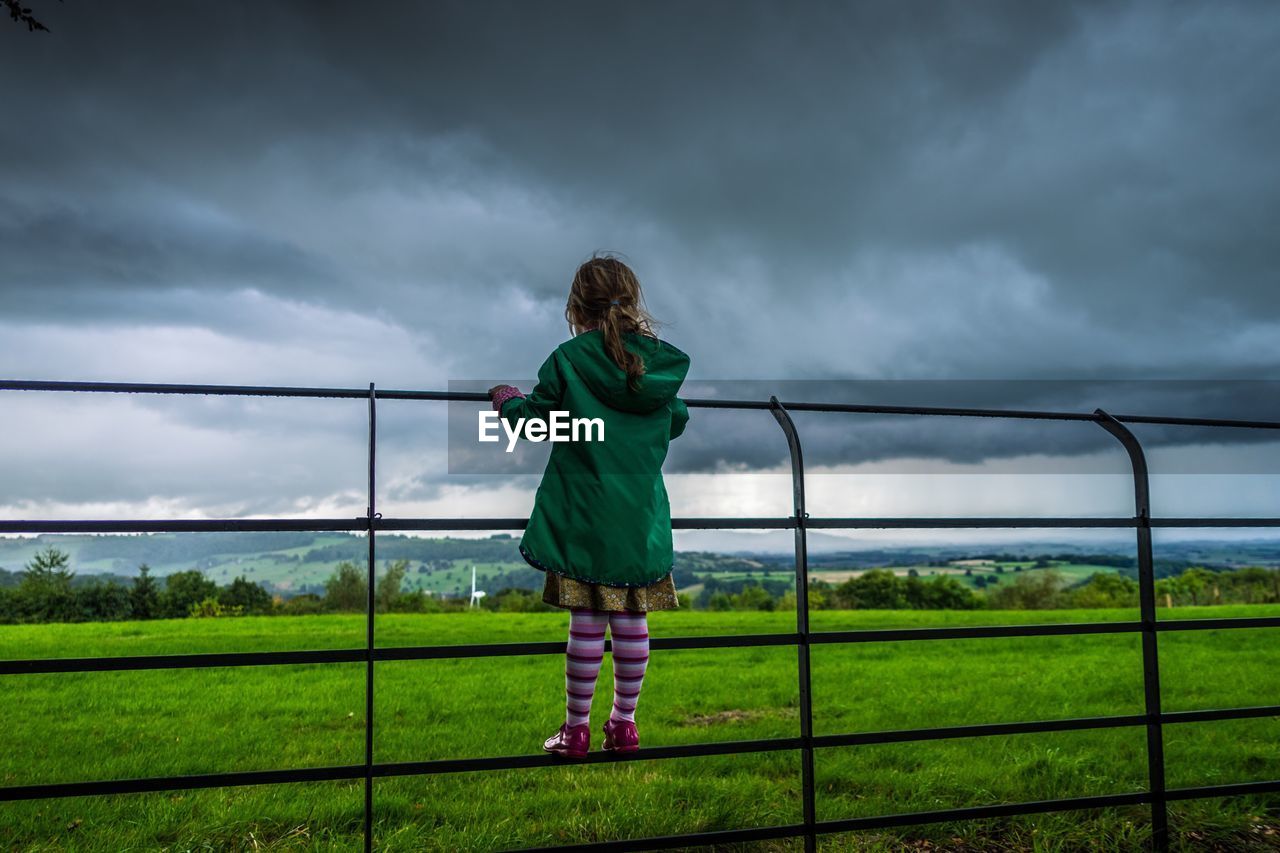 Rear view of girl standing on fence by grassy field against cloudy sky