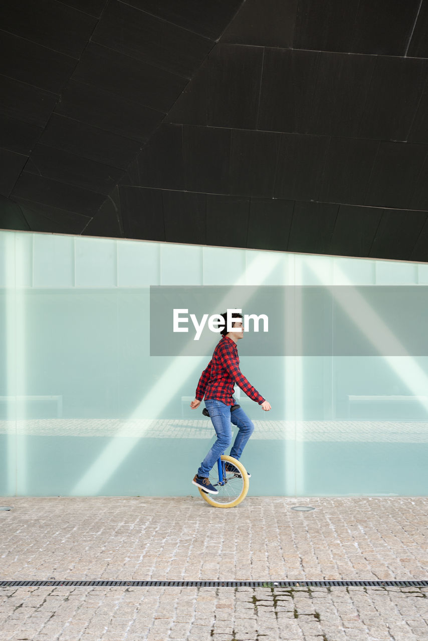 Full body side view of active young male in checkered shirt and jeans performing trick on unicycle near mirrored glass wall of contemporary building