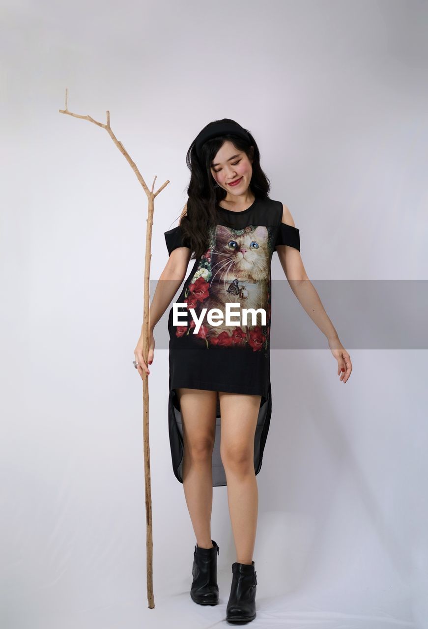 one person, full length, portrait, women, looking at camera, adult, studio shot, young adult, clothing, fashion, long hair, smiling, photo shoot, hairstyle, indoors, front view, happiness, footwear, shoe, human leg, limb, costume, gray, standing, emotion, black hair, gray background, high heels, dress, brown hair, female, white background, holding, black