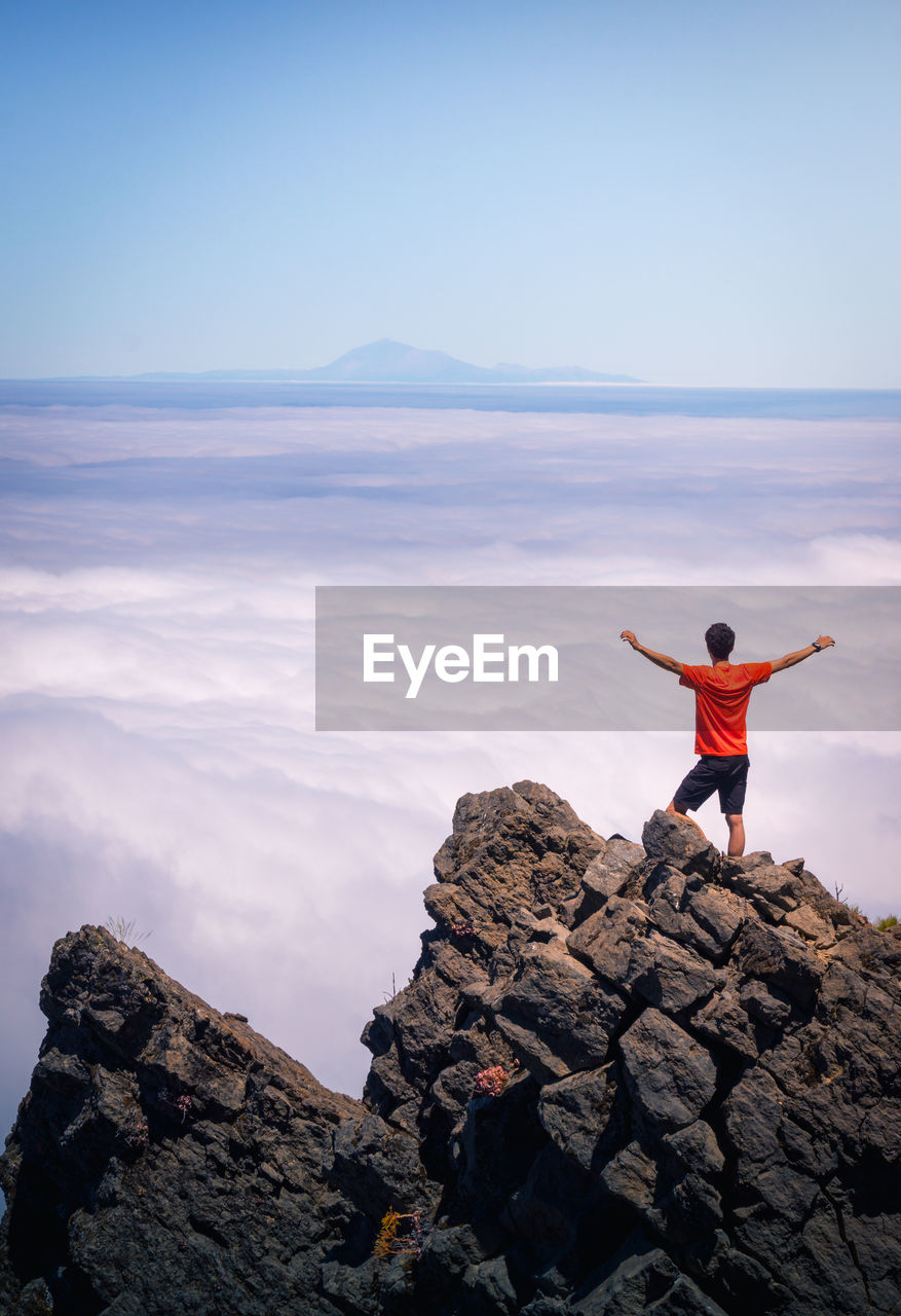 Back view of anonymous male traveler standing with outstretched arms on rocky hill in highlands and enjoying amazing view of clouds
