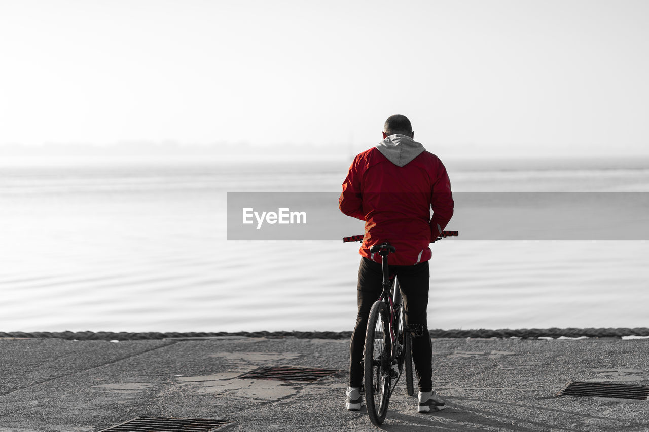 REAR VIEW OF MAN RIDING BICYCLE ON SHORE AGAINST SKY