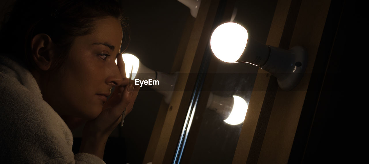 Young woman doing make-up in front of mirror with illuminated light bulbs in darkroom