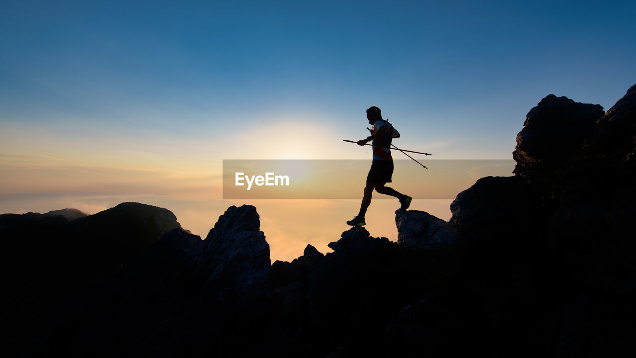 Sunset silhouette of skyrunner man descending from alpine ridge with poles