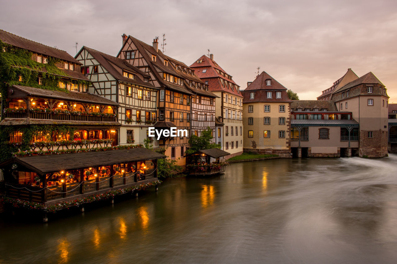 Illuminated buildings by river against sky in city at dusk