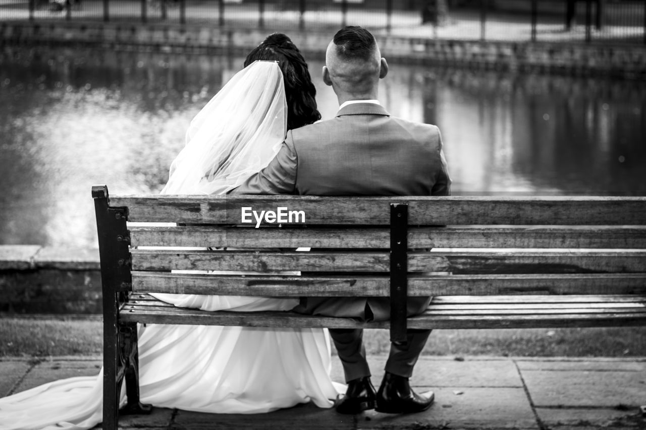 Rear view of wedding couple sitting on bench by lake