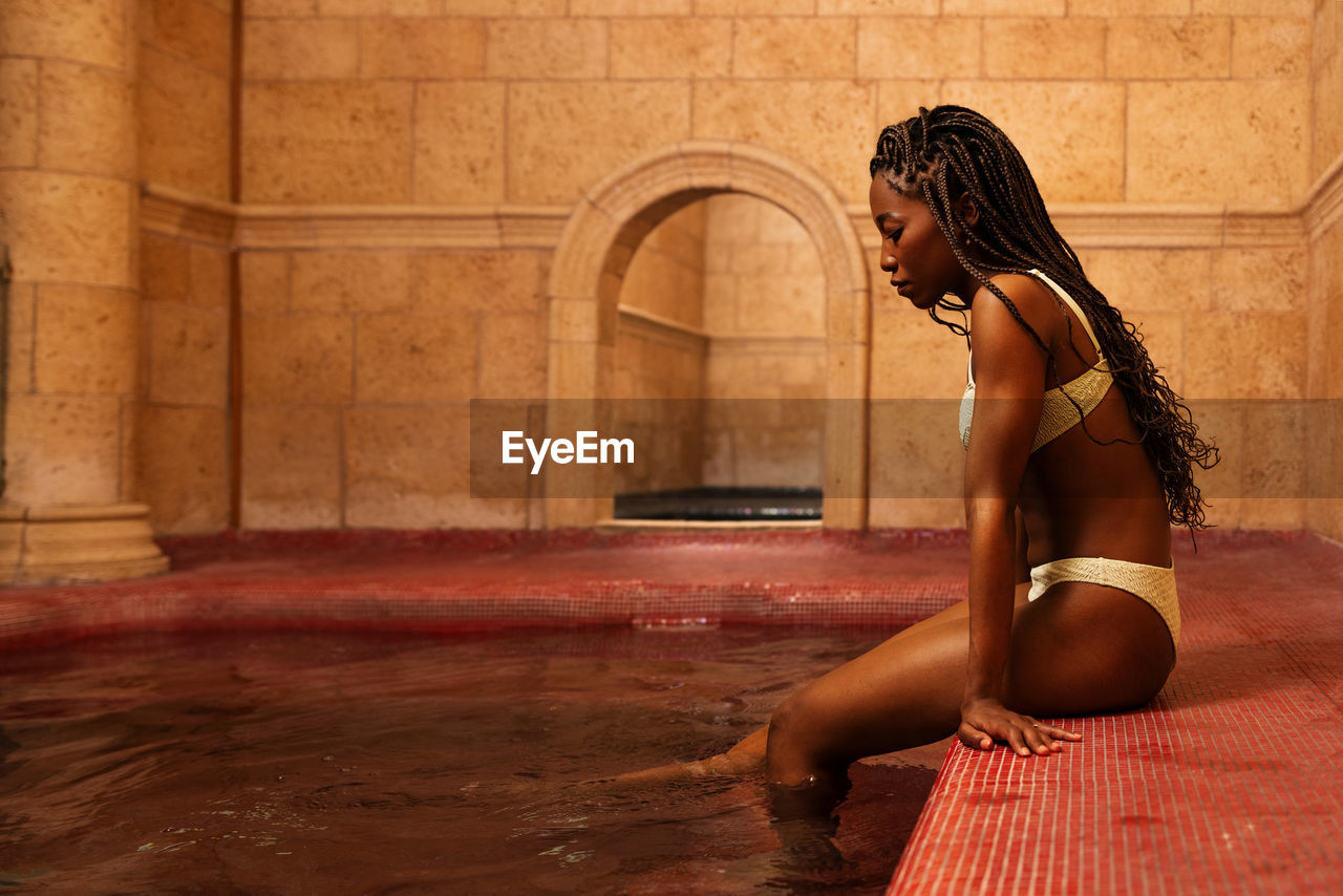 Side view of slim african american female in swimwear sitting with legs in water of pool while relaxing in spa center