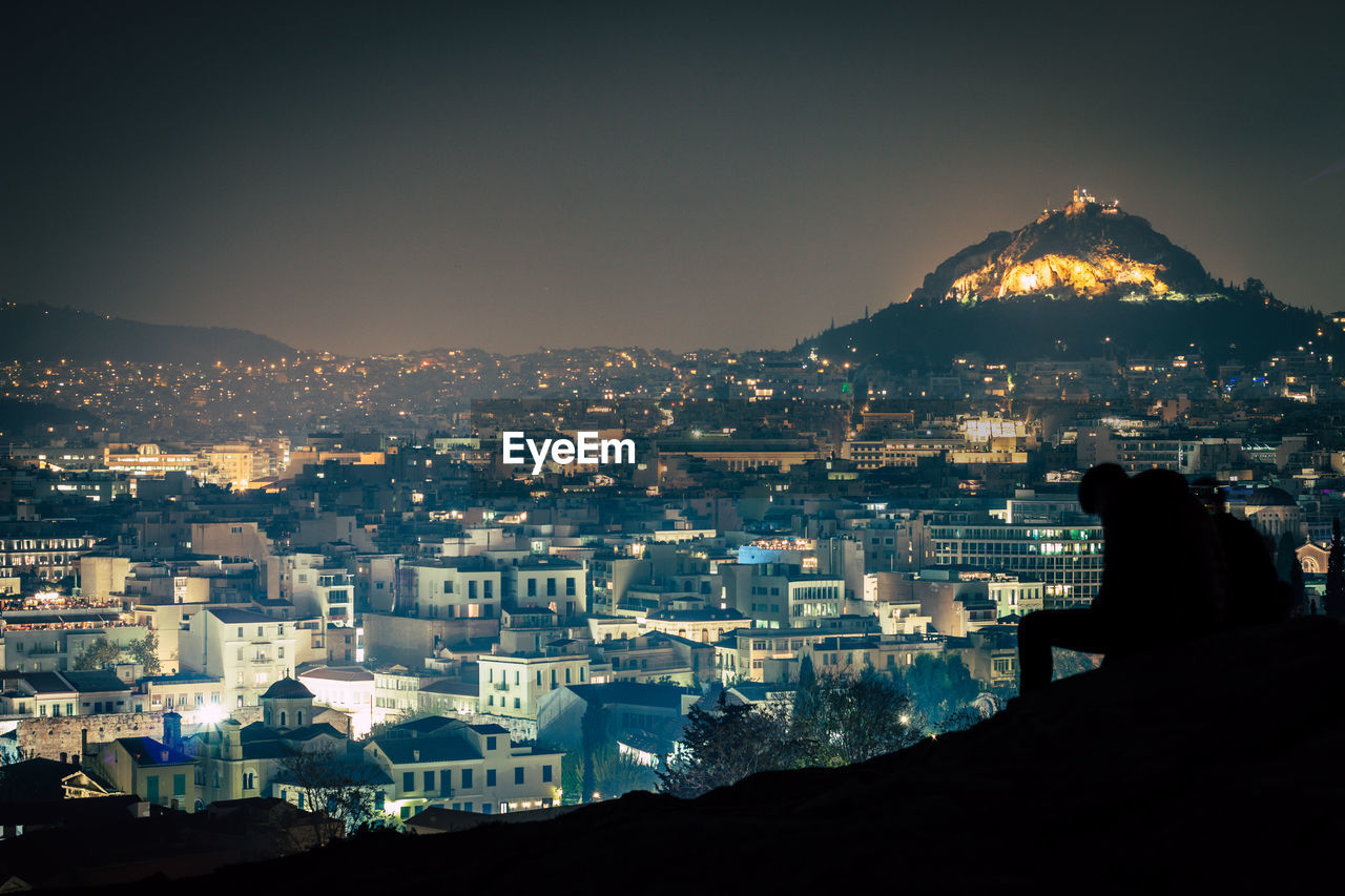 High angle view of illuminated cityscape with silhouette man sitting against sky