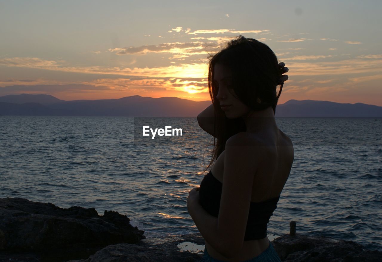 Side view of young woman standing at beach during sunset