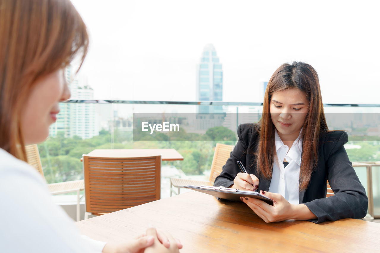 Businesswoman taking interview of woman sitting on desk against clear sky