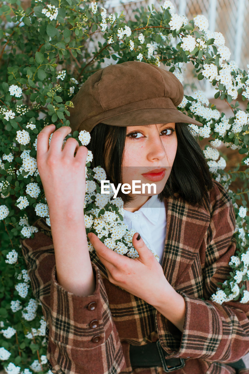 Young gentle ethnic female in checkered wear with beret touching blossoming plant while looking at camera on street