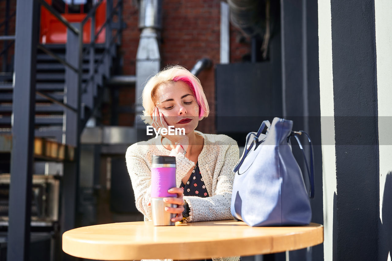 Serene informal female sitting at table with eco friendly cup with hot beverage in city on sunny day