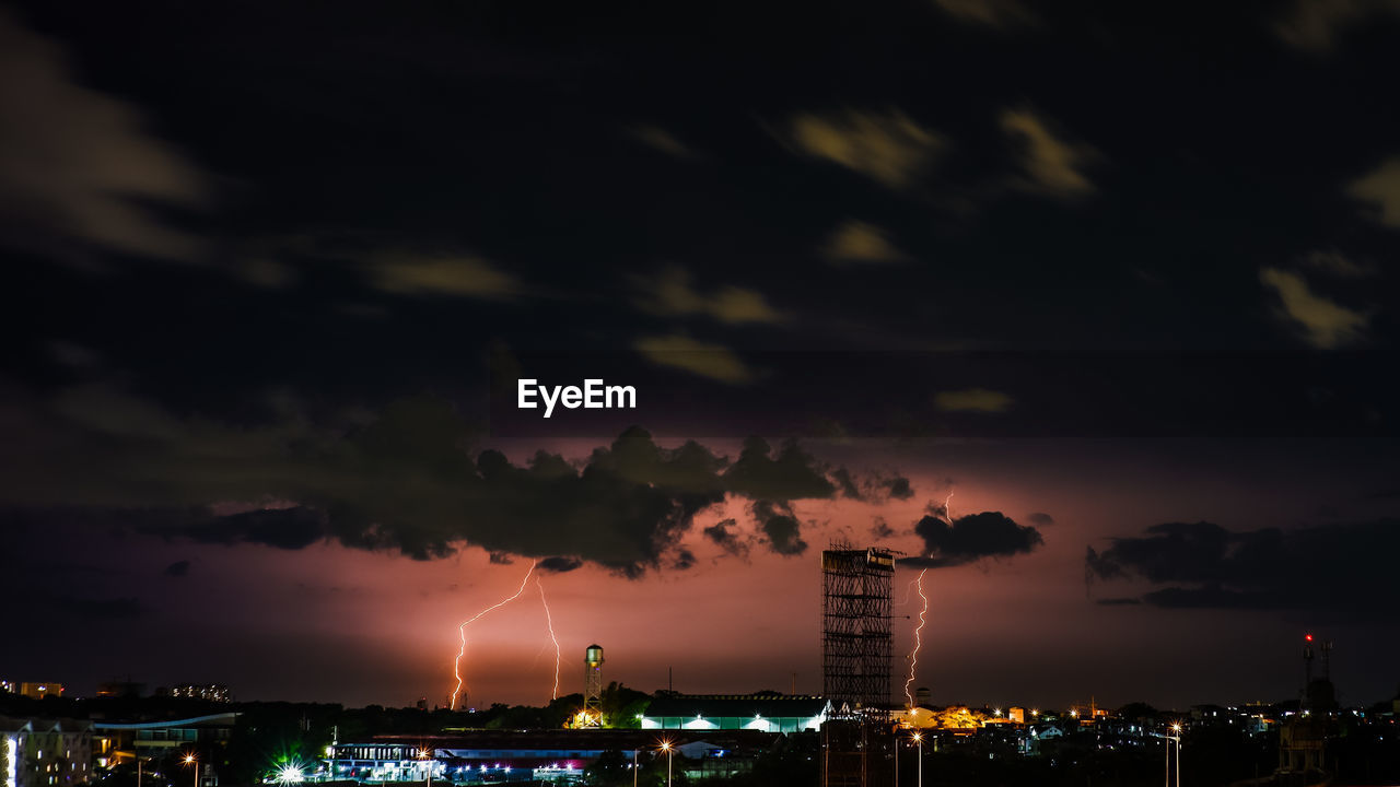 Illuminated buildings in city against dramatic sky at night
