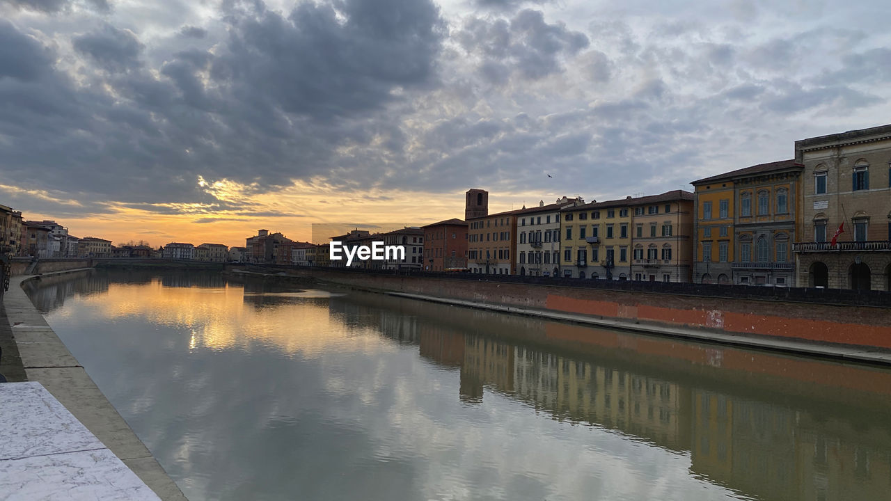 Buildings by river against sky during sunset