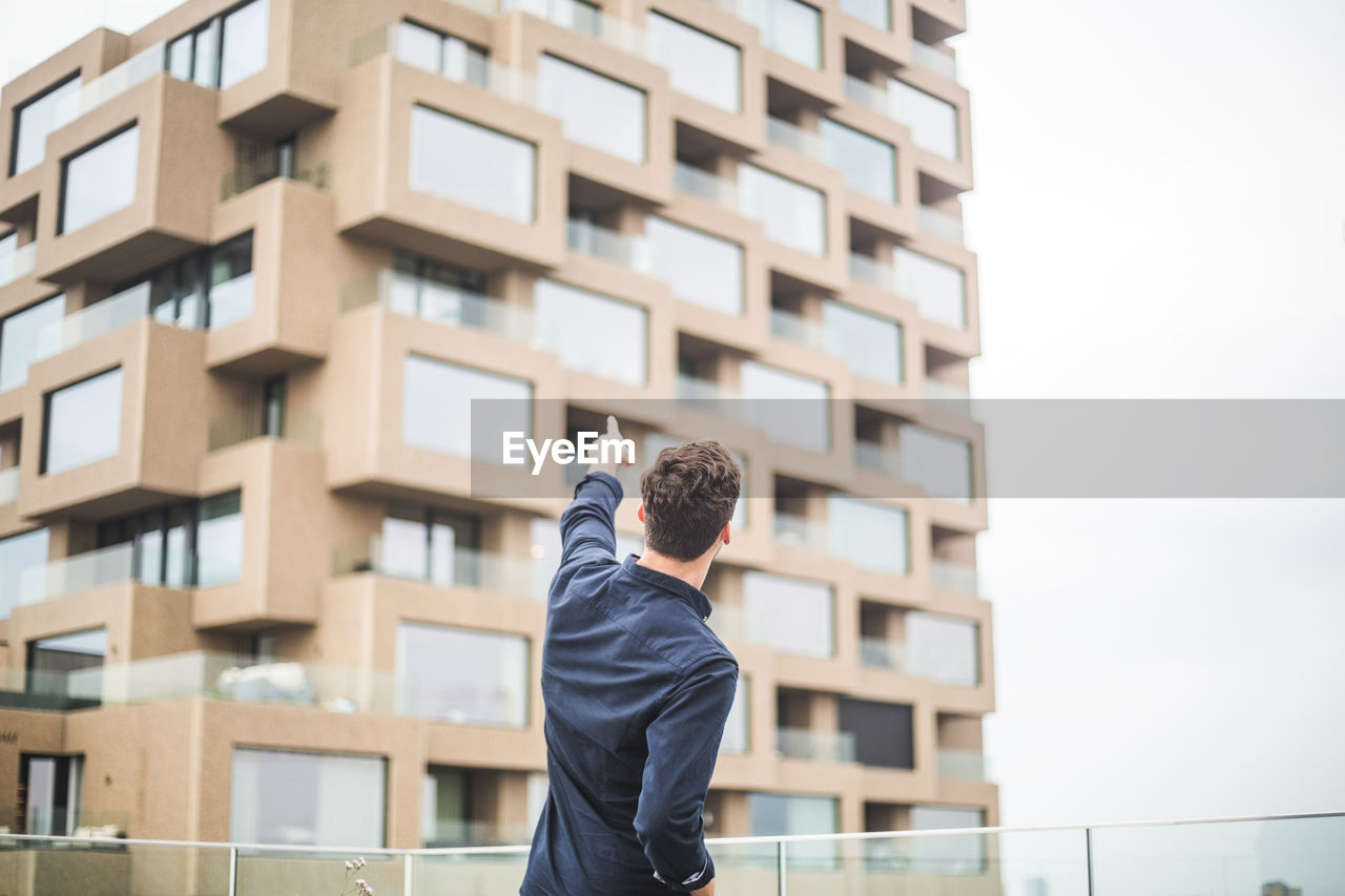 Rear view of man pointing at building while standing by glass railing on terrace