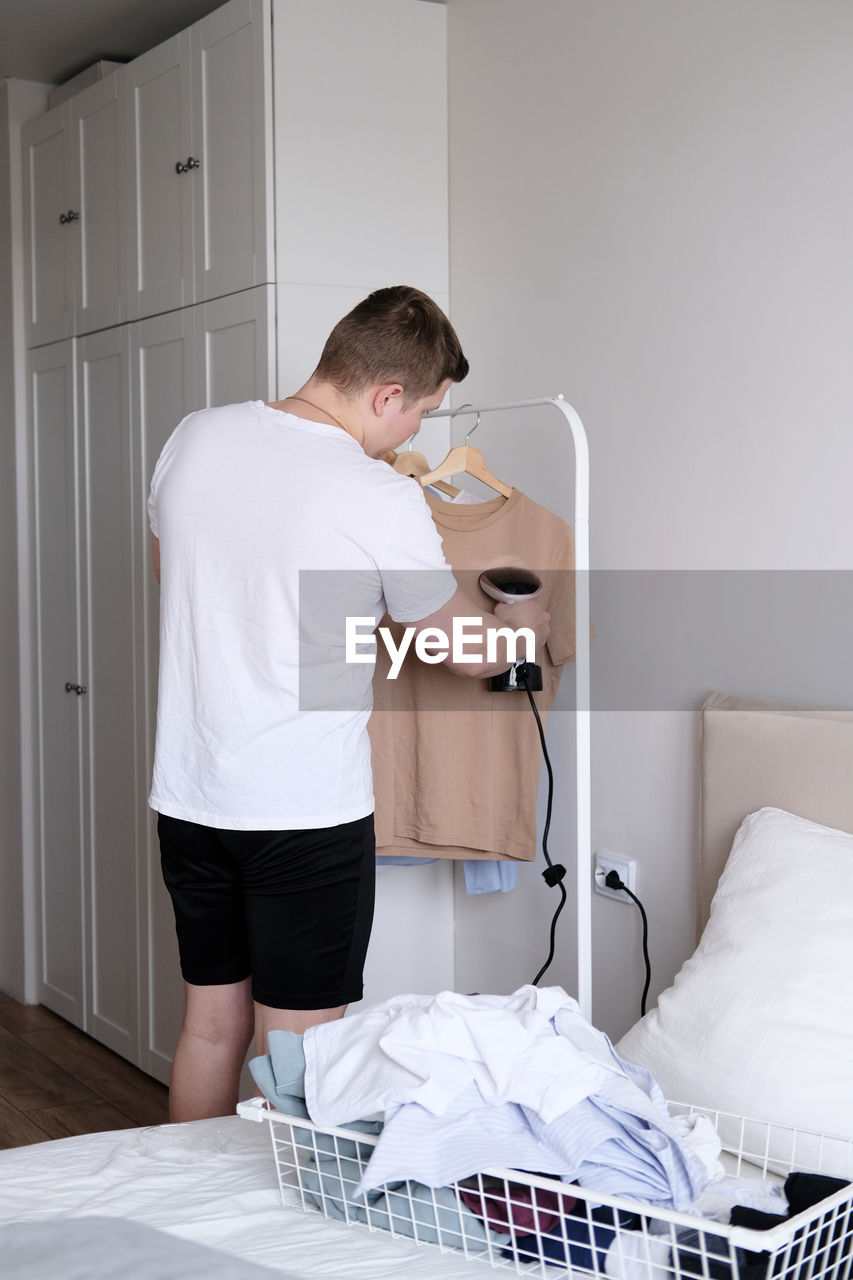 A young man holds the garment steamer in his hand and smoothes the t-shirt after washing and drying