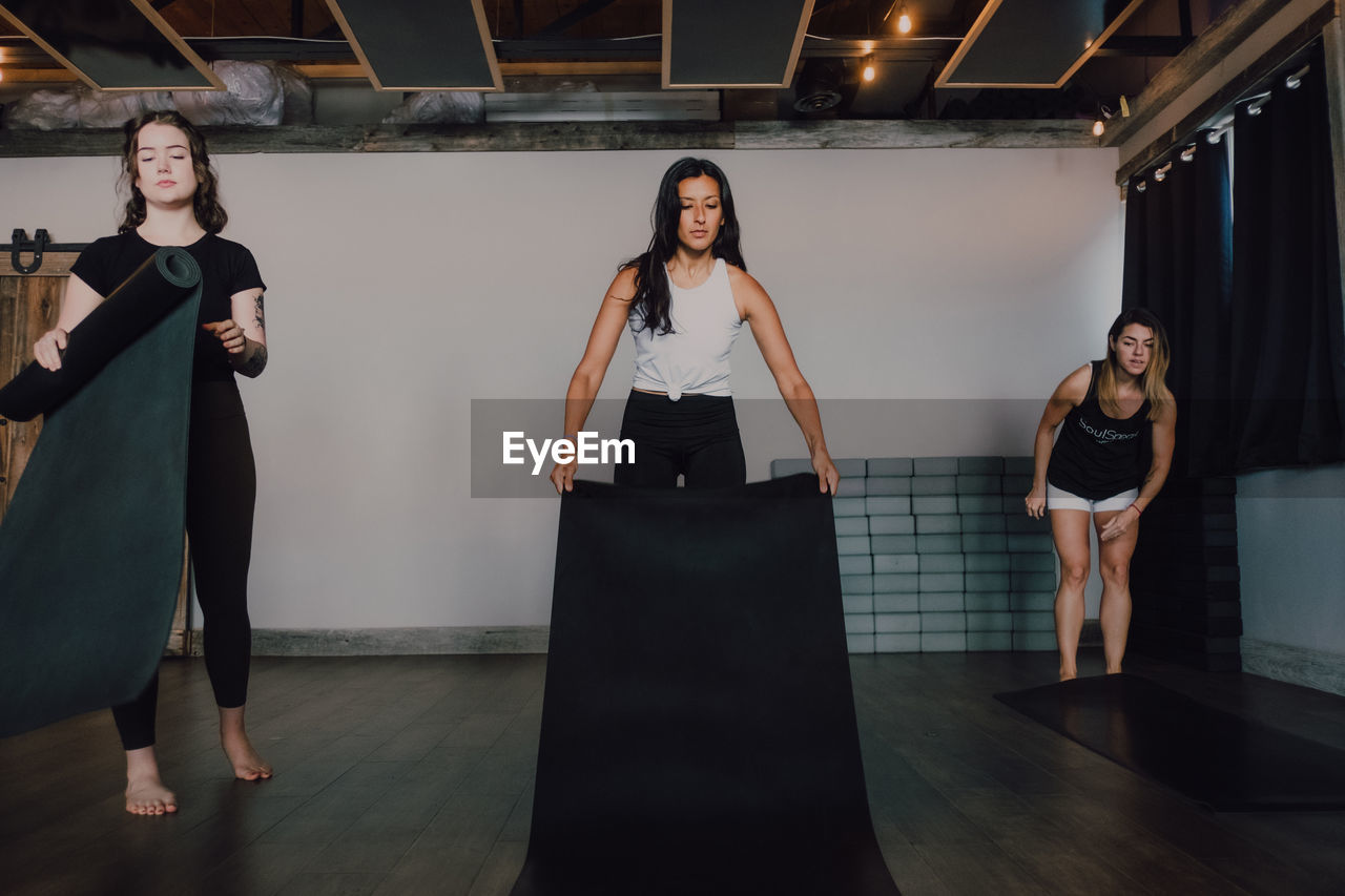 Low angle of calm female athletes in sportswear laying sports mats on wooden floor while preparing to train in spacious gym