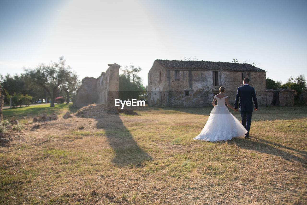 Full length rear view of newlywed couple walking by field by abandoned house against clear sky