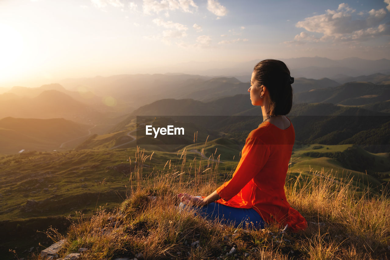 Young caucasian woman meditates in the lotus position in the grass in the mountains near the cliff