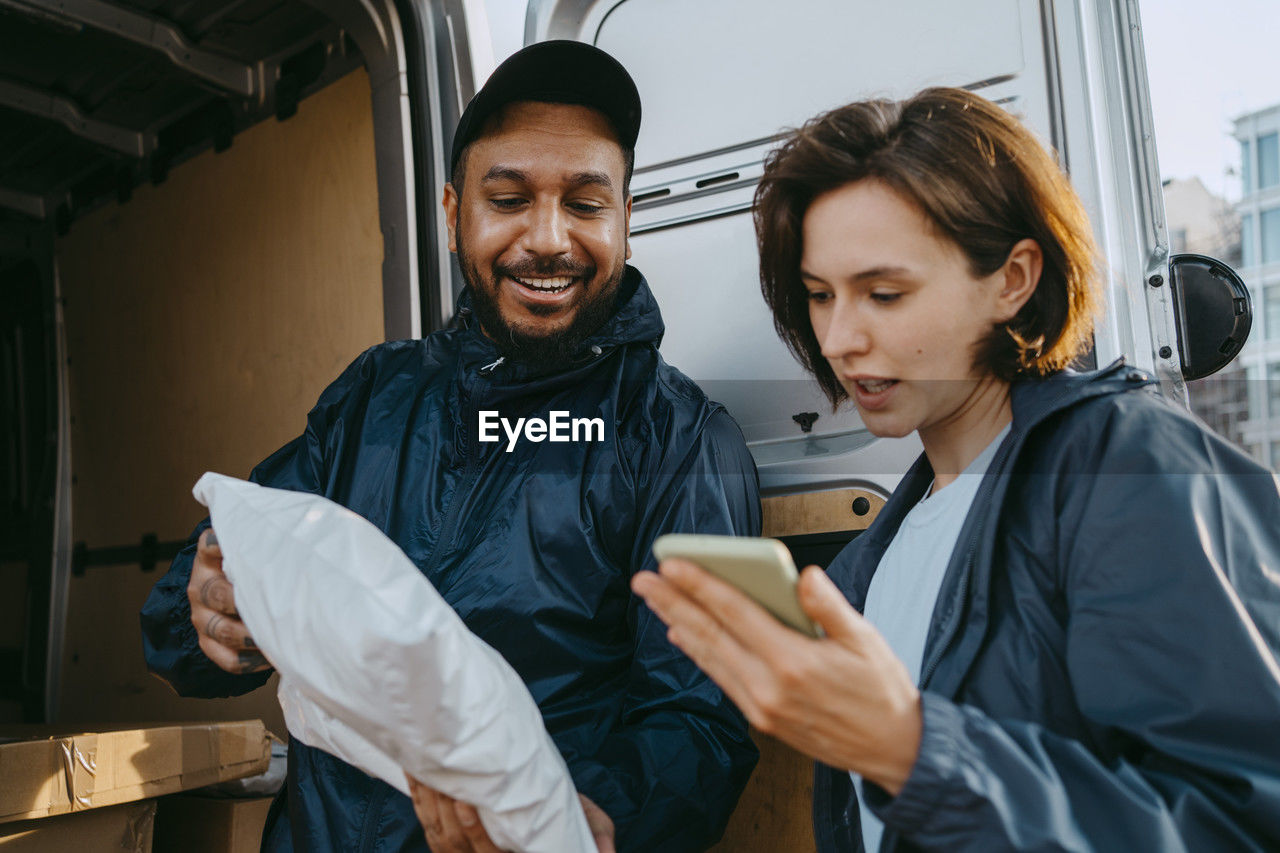 Smiling male delivery person showing parcel to female coworker holding smart phone near van