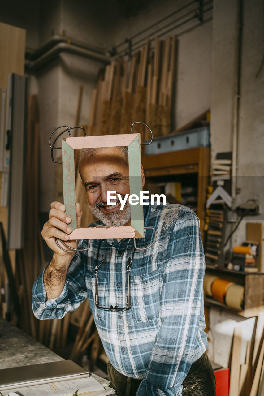 Portrait of happy craftsman looking through picture frame at workshop