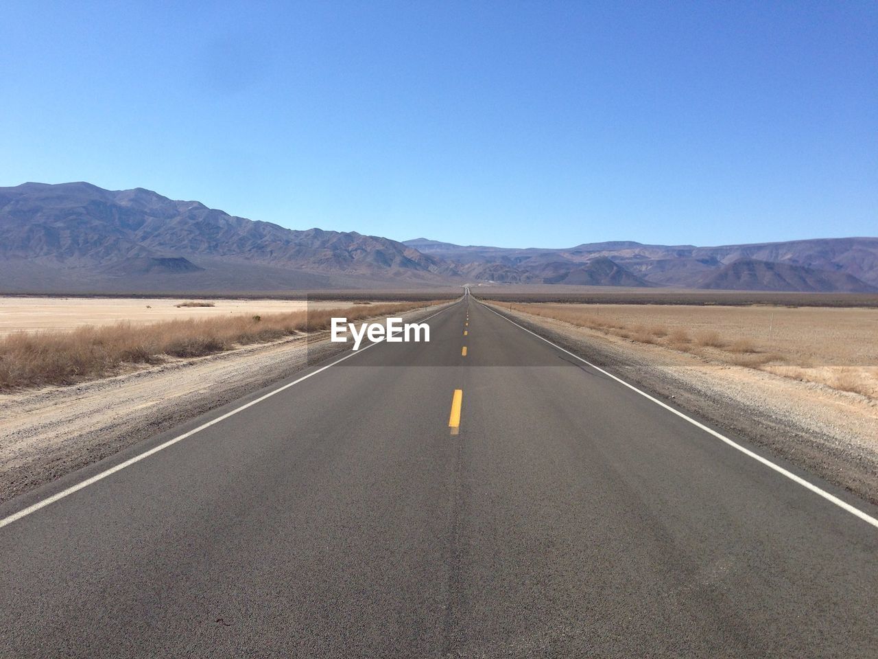 Empty road leading towards mountains against clear blue sky