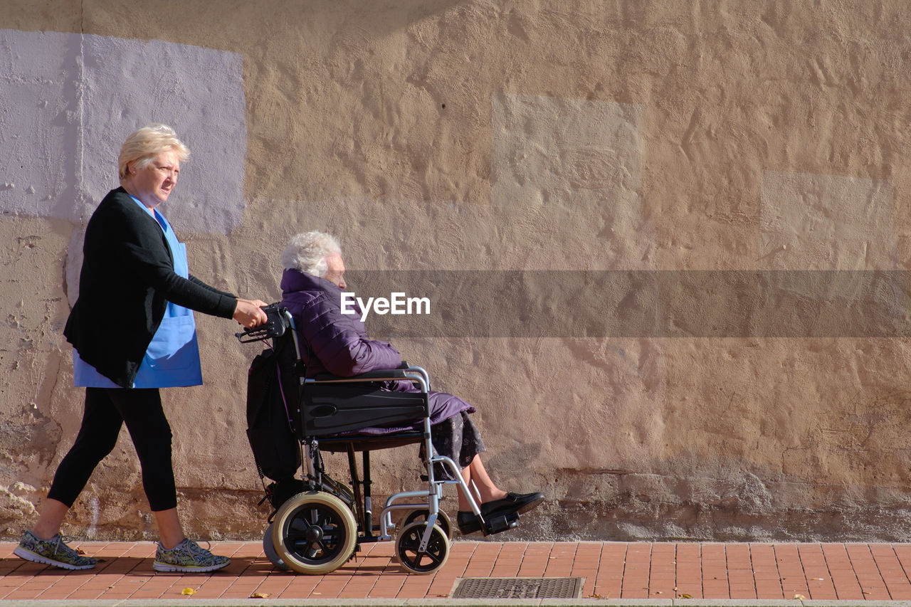 Caring nurse in uniform pushing elderly disabled woman in wheelchair while walking walking on paved sunny street near concrete wall in city