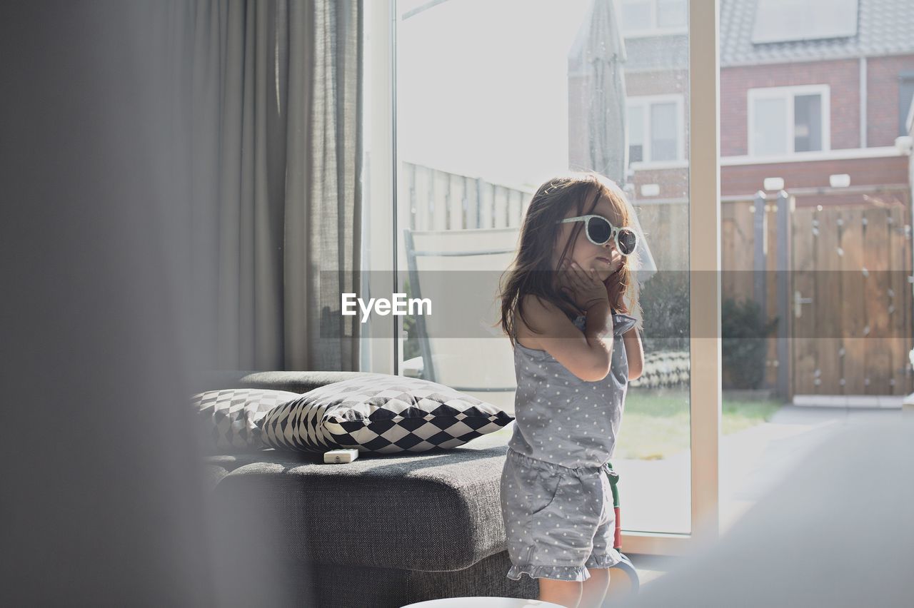 Girl wearing sunglasses while standing against window at home