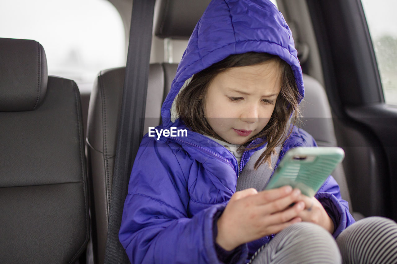 A little girl plays with phone in the backseat of a car on a car trip